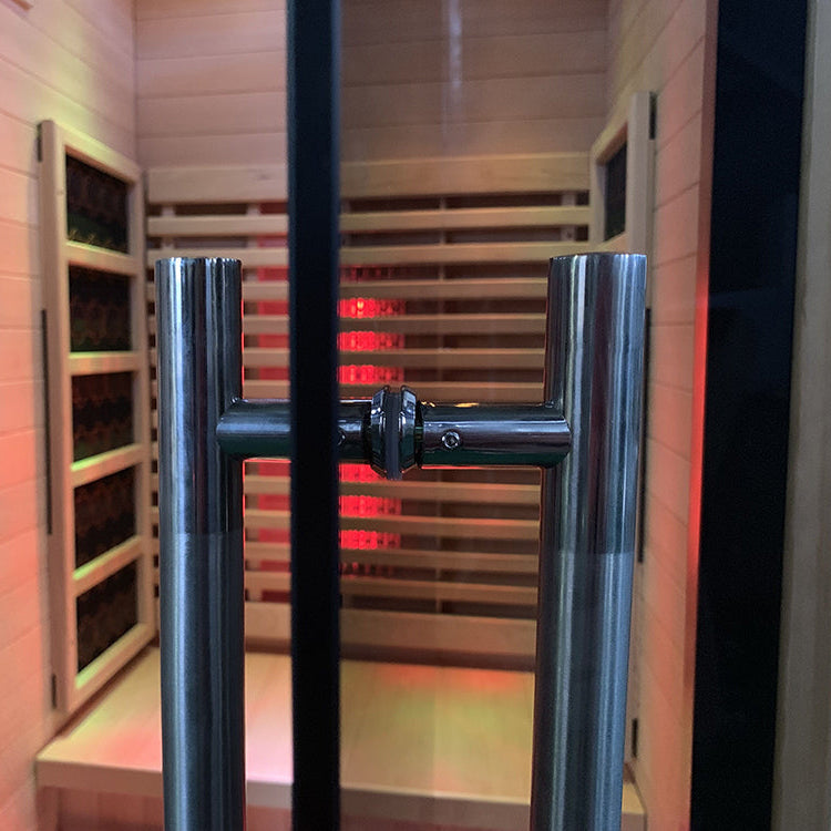 Lumin One Infrared Sauna interior view through glass door showing stainless metalic door handle white wood walls carbon heater panels glowing red on both sides and bench seat slats